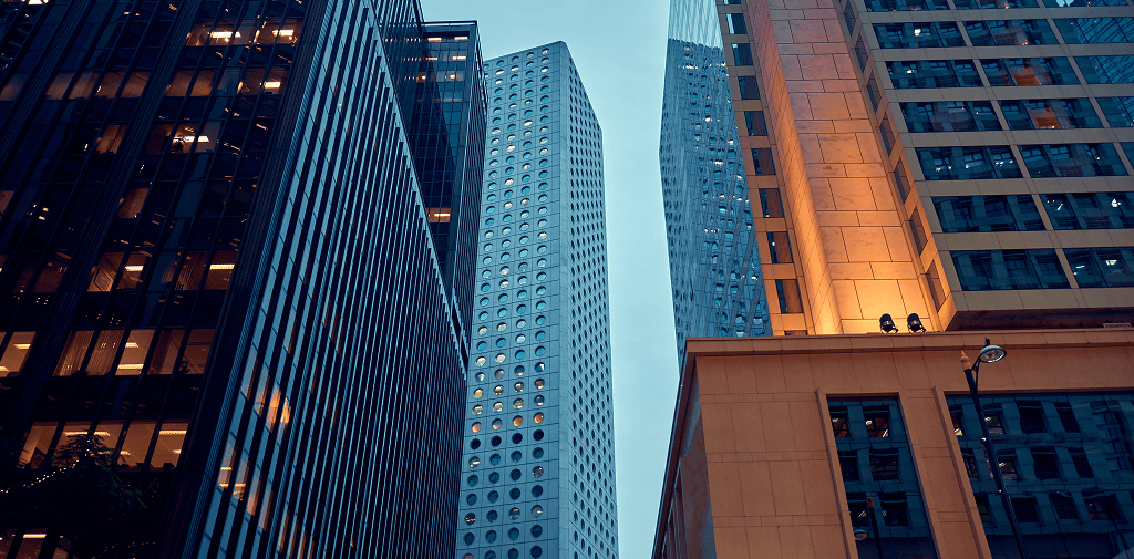 A low-angle view looking up at a cluster of modern city skyscrapers, highlighting a central tower with distinct circular windows surrounded by dark glass and warmly lit office buildings.